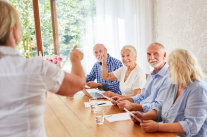 Referentin steht vor vier Senioren, die an Tisch nebeneinander sitzen 