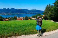 Person streckt die Hände seitlich nach oben, vor ihr Grasfläche, Häuser, See und Berge